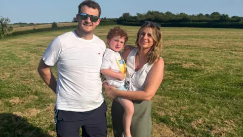 Richard stands with his family in a field on a sunny day. He has short dark hair and reflective sunglasses and a white t-shirt. A woman with blonde hair, her sunglasses on her head smiles beside him. She is holding a boy on her hip, with Richard leaning into his back. The boy has curly red hair. 