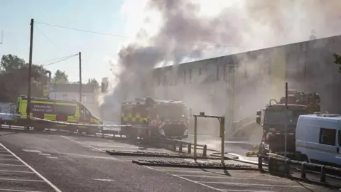 Jamie Lilley Fire engines and a van saying 'Enhanced rescue unit' can be seen outside the storage facility. Smoke billows out of the building.