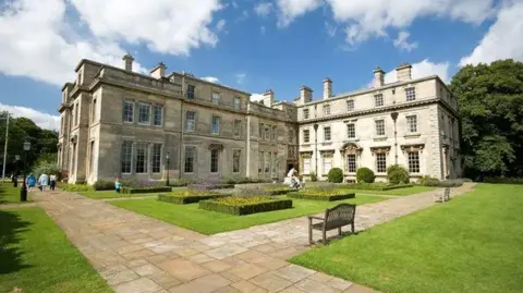 Normanby Hall The main building and a wing of a stately home, built in stone in the Regency style, with three storeys and tall windows. A formal garden includes raised flower beds, shrubs and sculptures. Wide paved paths surround the mansion and people can be seen walking past in the background. 