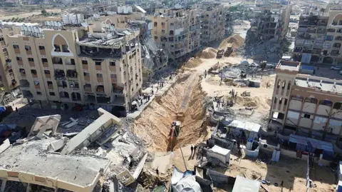 Anadolu via Getty Images Excavators dig a trench amid damaged and destroyed buildings in the Hamad City neighbourhood of Khan Younis (21 October 2025)