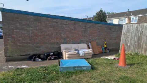 RSPCA Rubbish including a cream-coloured sofa, wooden furniture, orange traffic cone and a child's car seat abandoned next to a brick wall on a housing estate. 