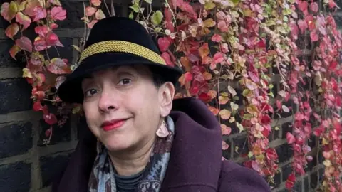 Natasha Gauthier Natasha pictured standing against a wall covered in red climbing plants wearing a purple coat and black hat with gold detailing and large earrings