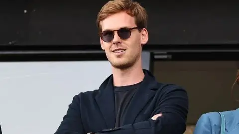 Getty Images Kyril Louis-Dreyfus with his arms crossed across his chest. He is smiling and has brown, short hair which is swept to the side. He is wearing round, dark sunglasses, a blue jacket and dark top underneath