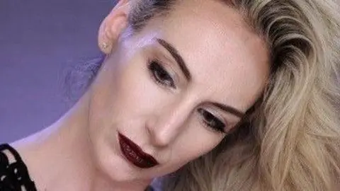 Instagram A blonde haired woman poses for the camera. She has her head tilted to the side and her hair is flipped over her head and dangling beneath her. She has dark make-up on, including deep red lips and black eyeliner. 