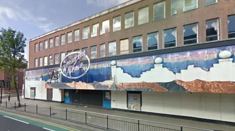 Google A mid-20th century, three-storey building, which looks as though it has seen better days. The ground floor features a gaudy facade including an evening skyline and the artistic outline of building tops. A large oval-shaped sign includes the words "Lexington Avenue" in a script-style font. The second and third floors have red-brick walls and long rows of windows. 