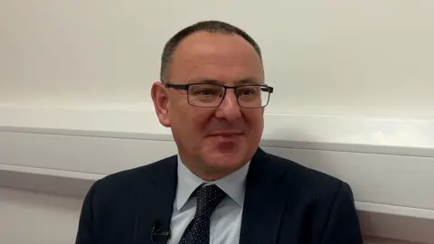 BBC Mike Cutland is smiling. He is wearing black rectangular glasses, a black suit with a blue shirt and black spotted tie. Behind him is a cream wall.