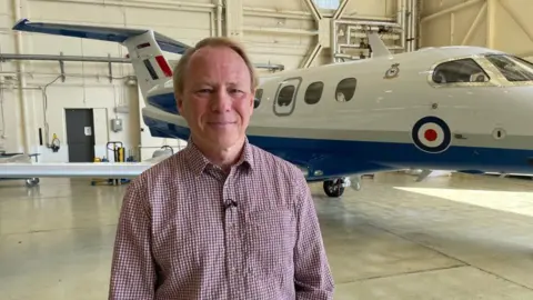 A man with short ginger hair and a long-sleeved red and white squared shirt is smiling into the camera. Behind him is a small aircraft which is white, blue and red, which is stationary inside a large warehouse building.