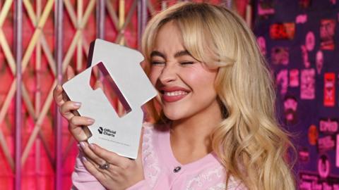 Sabrina Carpenter holding up her number one trophy from the Official Charts Company
