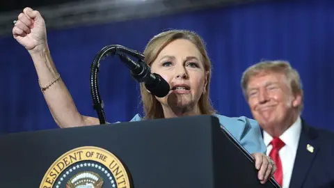 Darline Graham with her fist in the air as she gives a speech at a podium, with Trump looking on in the background