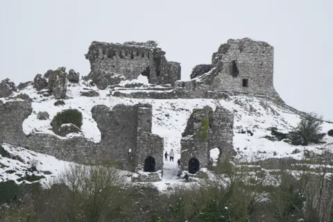 PA A ruined castle on a hilltop which is covered by snow. Walkers who are wrapped up warmly are walking among the ruins through the snow.