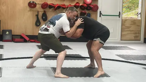Two men wrestling in a ring. The man on the left is wearing a white tshirt and green shorts, the other is all in black