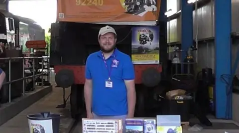 Parsons Family A man wearing a blue polo shirt stood in front of a train raised up off the ground.