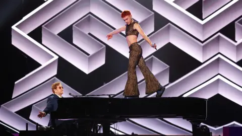 Getty Images Olly Alexander standing on top of a piano holding a microphone whilst Elton John plays the piano