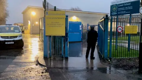 A police van is parked inside the gates of Kingsbury High School in Brent at dusk, while an officer stands nearby on a wet road with school buildings and signage visible.