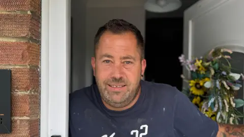 George King/BBC A head and shoulders image of Shaun Farrell. He is wearing a navy t-shirt and standing in his doorway. He is looking into the camera.
