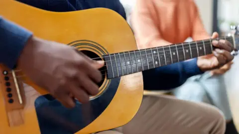 Close-up of man wearing a navy long-sleeved top playing an acoustic guitar with a female in a peach jumper in the background.