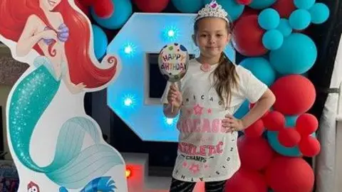 Merseyside Police Olivia stands in front of an LED number nine figure which is surrounded by red and blue balloons and a cardboard cut out of Princess Ariel. Olivia has her hand on her hip and is wearing a white t-shirt with pink wording and a crown.