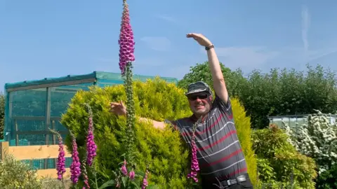 MWT A man with grey stubble and a beige sun shader hat wears a grey t-shirt and poses with his hands in the air in a garden with tall pink flowers, on a sunny day.