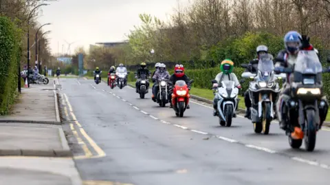 Roadrunnerpics A group of motorcyclists riding together in formation along a road. A few of the riders are wearing colourful novelty helmets or costumes, The road is bordered by trees and hedges.