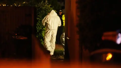 A man in a forensics suit walks towards a uniformed police officer.