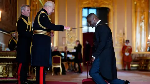 King Charles III dubs Sir Idris Elba with a sword on his shoulder. The King is wearing military dress uniform while Sir Idris is in a dark blue coat and blue trousers 
