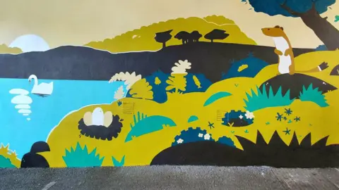 Falmouth BID Part of the mural which shows a stoat standing up on its back legs looking over a lake which has a swan on it. On the shore there is a nest with three large swan eggs and there are trees and bushes with flowers.
