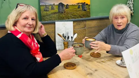 A picture of two ladies in a cafe with their cups of tea and coffee raised, looking at the camera.