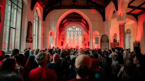 People standing in church which is being used as a music venue. There is a stage set up within a church alcove and a band is playing guitars. The stage is lit up with a red light.