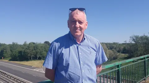 Lucas Vogan. He has short white hair, wearing sunglasses on his head and a light blue short sleeve shirt. He is standing on a motorway bridge with his hands behind his back.