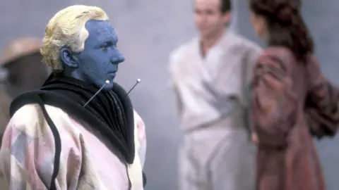 Dean Hollingsworth as Android in the "Doctor Who" sci-fi adventure "Timelash". He has blue skin and yellow hair.