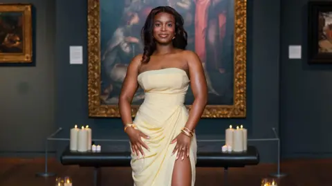 Emmanuel Dosumu/adidolvisuals A woman in a cream/yellow strapless dress smiles at the camera in front of a large painting.