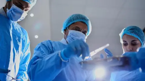 Three surgeons wearing face masks, gloves and blue scrubs look down at some surgical tools in preparation for an operation.