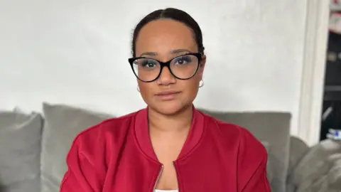 Shanice Aird, who is wearing black-rimmed glasses, a red jacket and has her dark hair tied back, she is sitting on a grey sofa