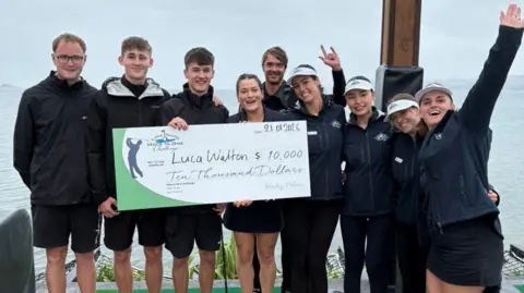 A group of people stood next to a lake. They are holding a large novelty cheque and smiling.