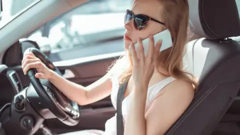 A woman is sitting behind the steering wheel with her seatbelt on, holding a mobile phone to her ear. She is looking at the road ahead and has one hand on the steering wheel.