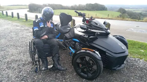 Jacob Davies-Pyke A man in a wheelchair, wearing black and blue motorcycle overalls and a black motorcycle helmet, next to a black motor trike parked by the side of the road. 