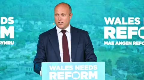 Getty Images Dan Thomas stands at a lectern with Reform branding