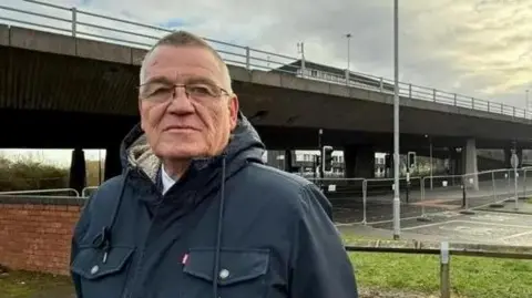 Martin Gannon standing in a car park near the Gateshead flyover. He has short hair and is wearing glasses and a navy coat. The concrete bridge is behind him. Temporary fencing has been put up across the road running under the bridge. 