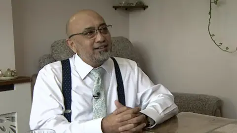 Nasar is wearing a white shirt with a green tie and glasses. He is sitting at a table with his hands clasped together and looks into the distance. 