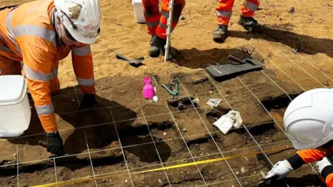 Two archaeologists wearing white hard hats and orange hi-vis suits on their knees at an excavation, taking samples from the ground. The soil is sandy coloured except on the corner of a rectangle over which they are bending. It is much darker in colour.