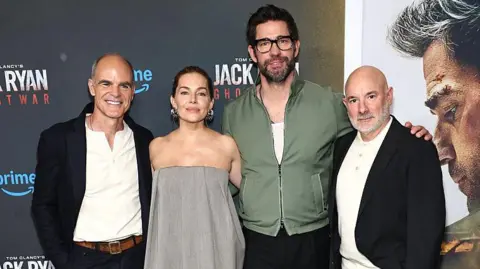 Getty Images Three men and a woman stand together in a line posing for photos during a film premiere. Two of the men are bald and wear similar black jackets with white tops underneath. The other man wears a green jacket with a white top underneath and glasses. The woman wears a strapless grey dress. 