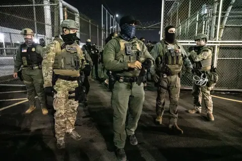 U.S. Immigration and Customs Enforcement (ICE) agents stand in front of chain link fence