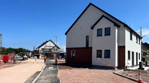 On the right of the photo is a semi-detatched property with a path leading up to it. On its left in the background is another property, surrounded by scaffolding, backed by a clear blue sky.