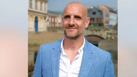 Businessman Austen Moore is wearing a light blue suit and white shirt. He is bald and has grey stubble. 