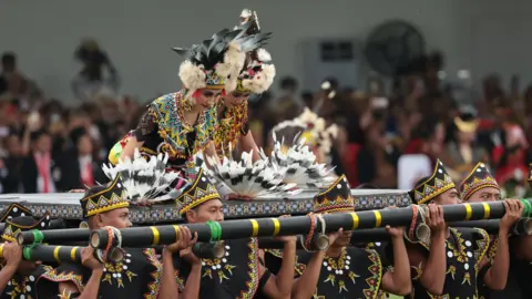 EPA Two dancers in costume sit upon a platform carried by men. 