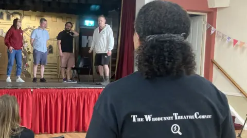 Back of a polo shirt showing the logo "The Whodunnit Theatre Company" with the stage in the background with members of the cast rehearsing