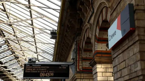 View inside Crewe railway station. There is a sign with the word Crewe on the right of the screen and a destination board for a train to London in the centre of the image.