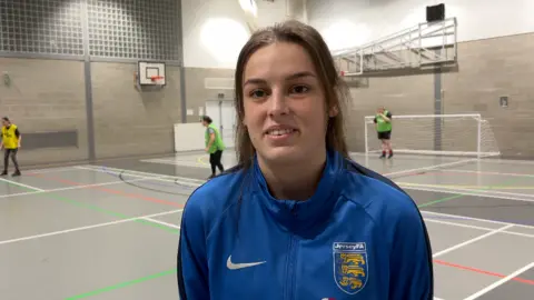 Charley has brown hair and a bright blue Jersey FA wind-breaker. She is stood on an indoor multi-purpose sports pitch that is grey with multi-coloured lines.