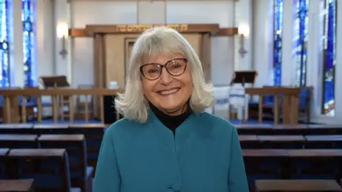 Shaun Whitmore/BBC A head and shoulders photo of Dr Marian Prinsley inside Norwich Synagogue. She is wearing a teal blue jacket, has mid-length grey/blonde hair and dark rimmed glasses. 