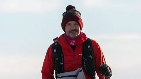  Mr Chamberlain wearing a red jacket and holding hiking sticks in his hands. He wears a red hat and a grey bag on his front. Behind him is the sky.